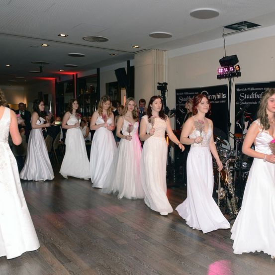 Eine Gruppe von Frauen in weißen Kleidern geht auf einer Tanzfläche, wahrscheinlich für eine Hochzeit. Hinter ihnen stehen Leute. Es gibt Musikinstrumente, Lautsprecher und Lichter an der Seite.