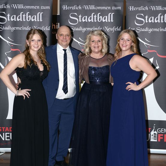 Vier Personen stehen lächelnd zusammen und posieren für ein Foto vor einem Banner, auf dem 'Herzlich Willkommen zum Stadtball' steht. Die Frau auf der linken Seite trägt ein schwarzes Kleid, während der Mann in der Mitte einen Anzug und eine Krawatte trägt. Die anderen beiden Frauen sind in blaue Kleider gekleidet.