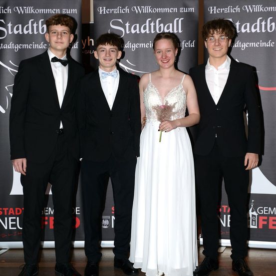 Vier junge Menschen stehen zusammen, schick gekleidet. Eine Frau in einem weißen Kleid steht zwischen zwei Männern in schwarzen Anzügen. Sie alle lächeln in die Kamera.