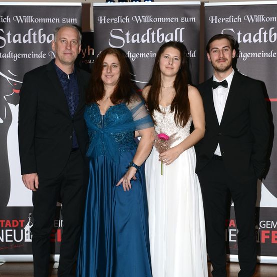 Eine vierköpfige Familie steht vor einem Banner für den Stadtball, mit zwei Frauen und zwei Männern, die formell gekleidet sind. Das Banner hat den Text 'Herzlich Willkommen zum Stadtball' im Hintergrund.