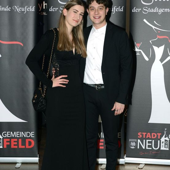 Ein junges Paar, eine Frau in einem schwarzen Kleid und ein Mann in einem schwarzen Anzug, posiert für ein Foto vor einem Hintergrund mit dem Text 'Stadt Neufeld'.