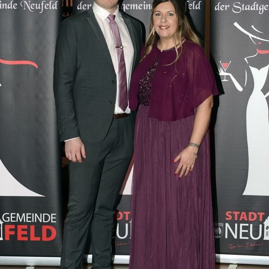 Ein Mann und eine Frau stehen nebeneinander, formell gekleidet, bei einer Veranstaltung. Sie lächeln und scheinen für ein Foto zu posieren. Hinter ihnen befindet sich ein Hintergrund mit Text und einer Zeichnung einer Frau, die ein Cocktailglas hält.