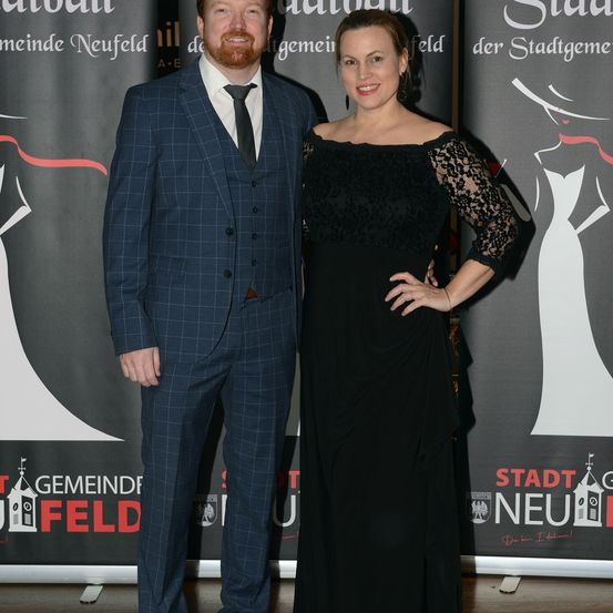 Ein Mann und eine Frau stehen lächelnd bei einer Veranstaltung zusammen. Der Mann ist in einem Anzug und die Frau in einem schwarzen Kleid. Sie stehen vor einer Kulisse mit dem Text 'Gemeinde Neufeld'.