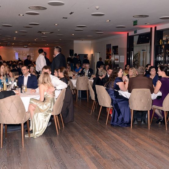 Ein Speisesaal mit vielen Menschen, die an Tischen sitzen, einige mit Gläsern. Einige stehen in der Nähe eines Bildschirms, andere in der Nähe einer Bar mit Flaschen. Der Raum hat Holzböden und eine Decke mit Lichtern.