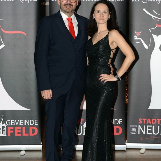 Ein Mann und eine Frau stehen gemeinsam auf einem roten Teppich und tragen formelle Kleidung. Der Mann trägt einen Anzug und eine Krawatte, während die Frau ein schwarzes Kleid trägt. Sie posieren für ein Foto. Hinter ihnen befinden sich Banner mit Text und Bildern.