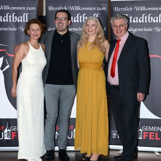 Vier Personen posieren für ein Foto. Sie stehen vor einem Hintergrund mit deutschem Text. Die Frau im weißen Kleid und die Frau im gelben Kleid lächeln.