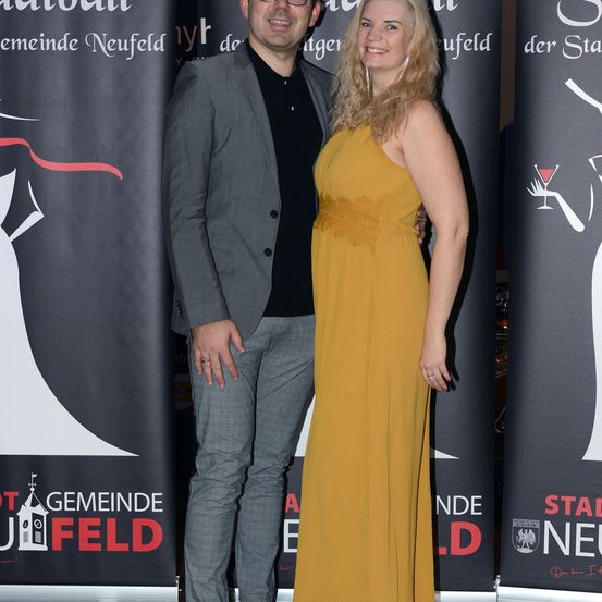 Ein Mann in einem grauen Anzug und eine Frau in einem gelben Kleid stehen lächelnd vor einem Hintergrund mit Text.