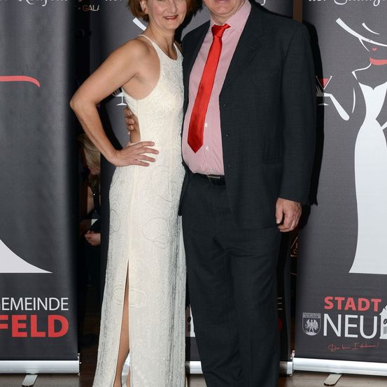 Ein Mann und eine Frau stehen eng beieinander, formell gekleidet, vor einem Hintergrund mit dem Text 'Stadt Neu'.