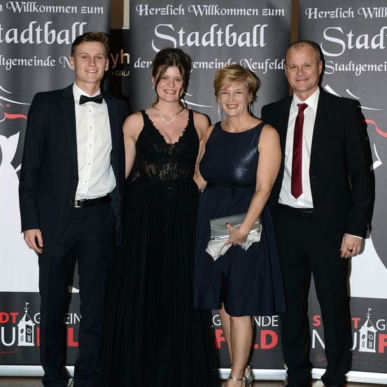 Vier Menschen in formeller Kleidung, zwei Frauen und zwei Männer, stehen zusammen, lächeln und posieren für ein Foto. Hinter ihnen ist ein Hintergrund mit dem Text 'Herzlich Willkommen zum Stadtball'.