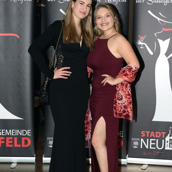 Zwei Frauen stehen nebeneinander bei einer Veranstaltung, lächeln und posieren für ein Foto. Sie sind in Abendkleidern gekleidet, eines in Schwarz und das andere in Weinrot. Hinter ihnen befinden sich Banner mit Logos und Text.