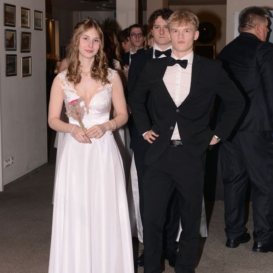 Eine junge Frau in einem weißen Kleid und ein junger Mann in einem schwarzen Frack stehen mit anderen in einem Korridor bei einer formellen Veranstaltung.