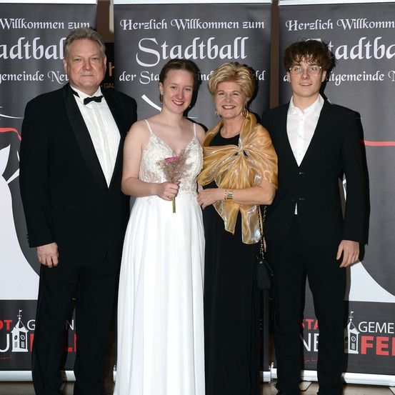 Vier Personen stehen vor einem Hintergrund, auf dem 'Herzlich Willkommen zum Stadtball' steht. Zwei Männer in Anzügen, eine Frau in einem schwarzen Kleid und eine Frau in einem weißen Kleid, die eine Blume hält.