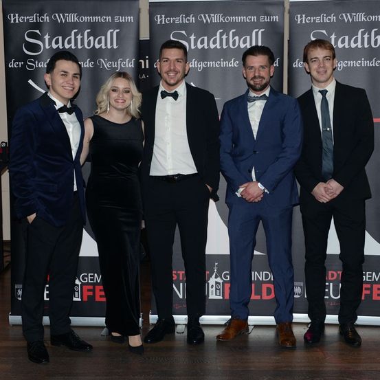 Fünf Personen in formeller Kleidung, einschließlich Anzüge und Krawatten, stehen vor einem Hintergrund mit der Aufschrift 'Stadtball'.