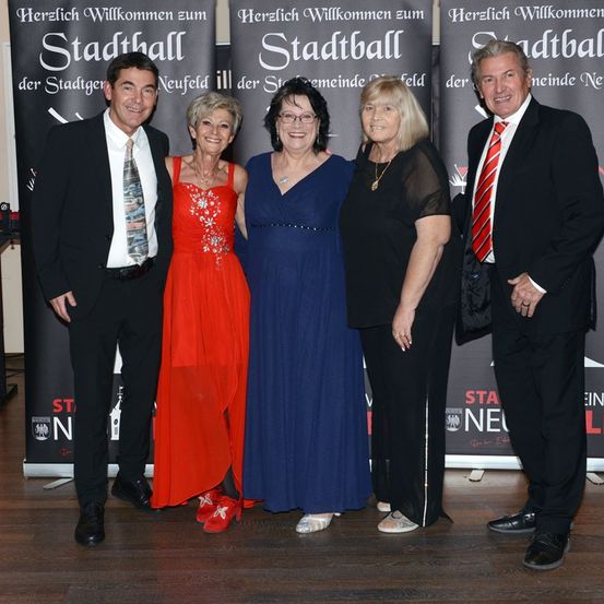 Fünf Personen stehen zusammen vor einem Banner. Zwei Männer und drei Frauen, sie sind alle formell gekleidet. Die Frauen tragen Kleider, eines in Rot, eines in Blau und eines in Schwarz. Die Männer sind in Anzügen. Das Banner hinter ihnen hat Text auf Deutsch.