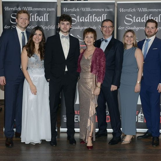 Eine Gruppe von sieben Menschen, bestehend aus Männern und Frauen, steht gemeinsam für ein Foto. Sie sind alle feierlich gekleidet. Hinter ihnen befinden sich Banner mit dem Text 'Herzlich Willkommen zum Stadtball der Stadtgemeinde Neufeld'.