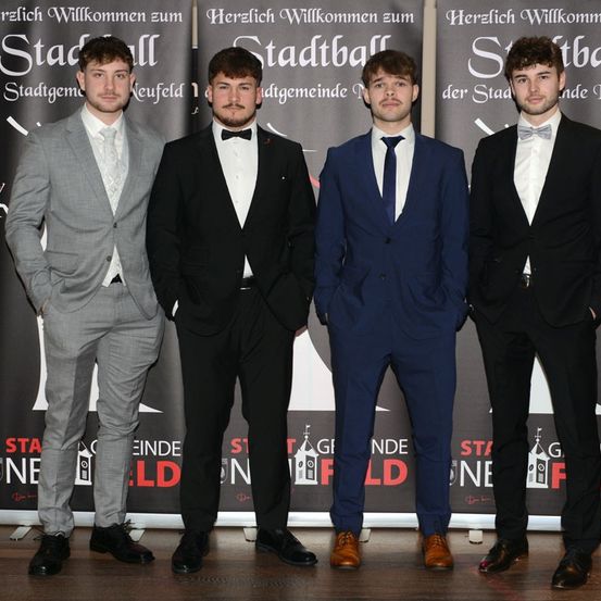 Fünf junge Männer, alle schick gekleidet, stehen zusammen vor einem Hintergrund mit Text und Logos und lächeln für ein Foto.
