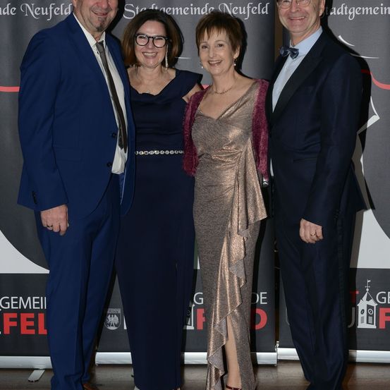 Vier Menschen stehen eng beieinander und lächeln für ein Foto bei einer Veranstaltung. Zwei Frauen tragen formelle Kleider, eine mit einer Halskette, und zwei Männer in Anzügen. Dahinter ist eine Kulisse mit deutschem Text.