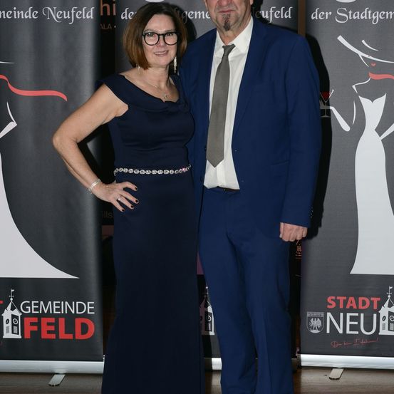 Ein Mann und eine Frau in formeller Kleidung stehen nebeneinander bei einer Veranstaltung, mit Bannern dahinter.