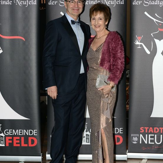 Ein Mann und eine Frau in formeller Kleidung stehen lächelnd vor einem Banner mit dem Text 'Der Stadt' und 'Gemeind Neufeld'.