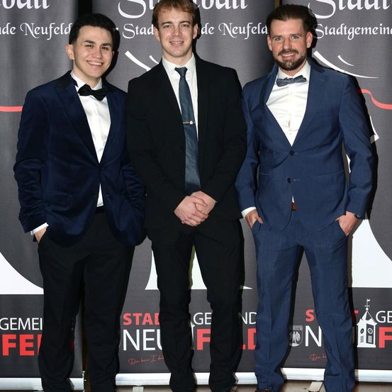 Drei Männer in formeller Kleidung posieren für ein Foto vor einem Hintergrund. Sie lächeln und stehen eng beieinander. Der Mann auf der linken Seite trägt einen Samtanzug, während die anderen beiden Anzüge mit Fliegen tragen.