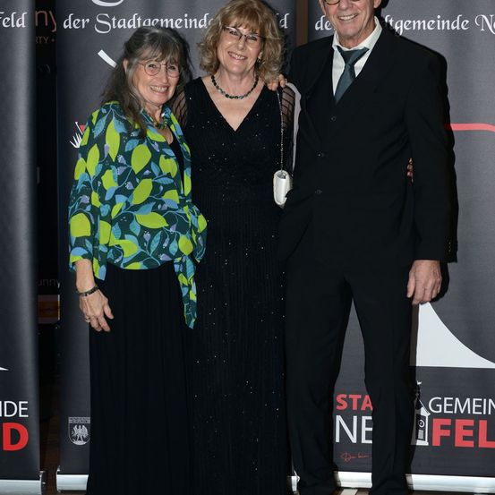 Drei Menschen, möglicherweise eine Familie, stehen eng beieinander. Die Frau links trägt ein grün-blau gemustertes Oberteil, die Frau in der Mitte ein schwarzes Kleid und der Mann einen schwarzen Anzug mit Krawatte. Sie alle lächeln.