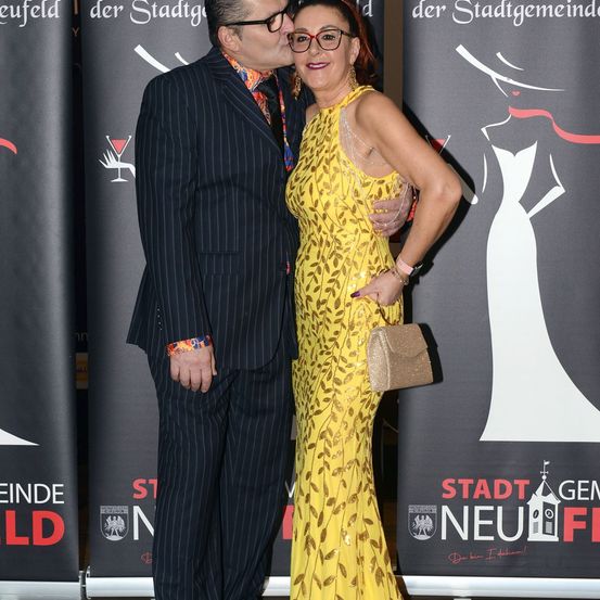 Ein Mann und eine Frau stehen bei einer Veranstaltung eng beieinander. Der Mann trägt einen schwarzen Anzug, und die Frau trägt ein gelbes Kleid. Hinter ihnen ist ein Banner mit den Worten 'Stadt Neu Fi.'
