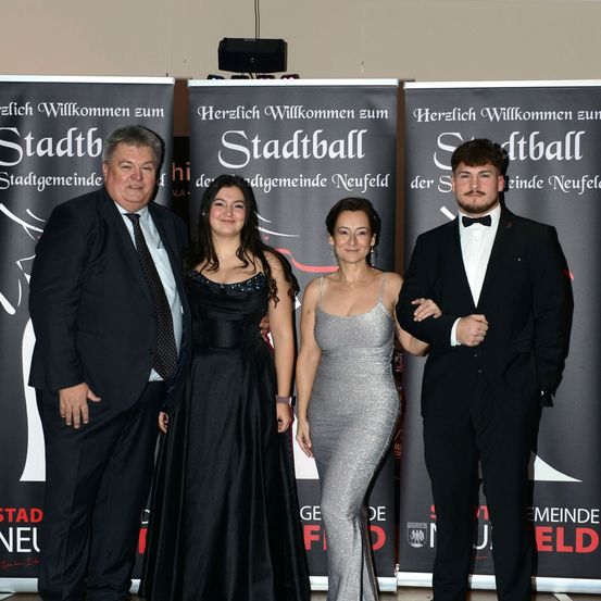 Vier Menschen in formeller Kleidung stehen vor Bannern mit dem Text 'Herzlich Willkommen zum Stadtball'. Sie lächeln und posieren für ein Foto.