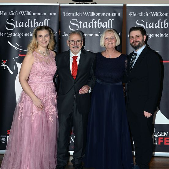 Vier Personen stehen eng beieinander und posieren für ein Foto bei einer Veranstaltung. Die Frau auf der linken Seite trägt ein rosa Kleid und eine Halskette. Der Mann in der Mitte trägt einen schwarzen Anzug mit roter Krawatte. Die Frau auf der rechten Seite trägt ein blaues Kleid. Der Mann auf der rechten Seite trägt einen schwarzen Anzug mit gestreifter Krawatte.