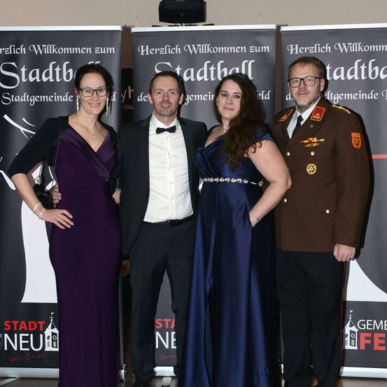 Vier Personen stehen für ein Foto bei einer Veranstaltung zusammen. Sie lächeln und schauen in die Kamera. Hinter ihnen sind Banner mit Texten und Logos.