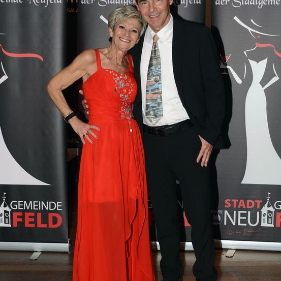 Ein Mann und eine Frau stehen eng beieinander, wahrscheinlich bei einer Veranstaltung. Die Frau trägt ein rotes Kleid und der Mann einen Anzug mit gemusterter Krawatte. Sie lächeln und posieren für ein Foto. Hinter ihnen befindet sich ein Hintergrund mit Text und Bildern.