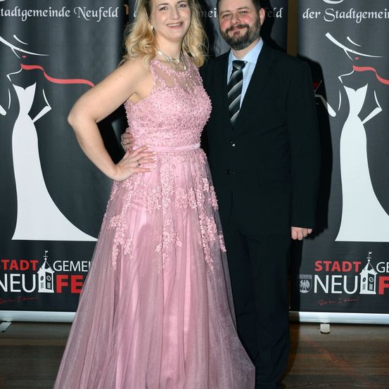 Ein Mann und eine Frau stehen zusammen, wahrscheinlich bei einer Veranstaltung. Die Frau trägt ein rosa Spitzenkleid und der Mann ist in einem schwarzen Anzug mit einem gestreiften Krawatte. Hinter ihnen ist ein schwarzer Hintergrund mit den Worten Stadt Neufeld.