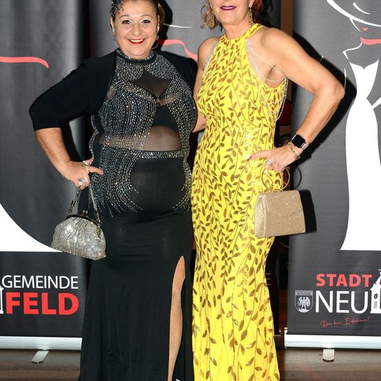 Zwei Frauen stehen bei einer Veranstaltung nebeneinander und lächeln. Die Frau auf der linken Seite trägt ein schwarzes Kleid mit Mesh-Oberteil. Die Frau auf der rechten Seite trägt ein gelbes Kleid mit Blattmuster.