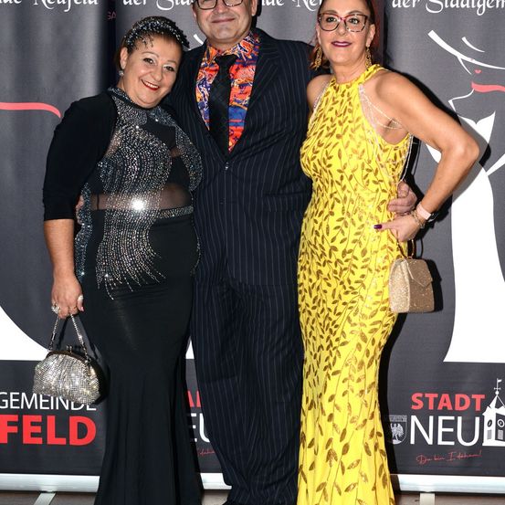 Drei Personen posieren für ein Bild auf dem roten Teppich. Eine Frau in einem schwarzen Kleid und eine andere in einem gelben Kleid stehen neben einem Mann in einem Anzug. Sie lächeln und halten Handtaschen. Dahinter befindet sich ein Hintergrund mit Text.