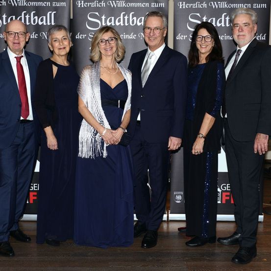 Sechs Menschen stehen zusammen auf einem Holzboden, wahrscheinlich bei einer Veranstaltung. Sie tragen alle formelle Kleidung. Drei von ihnen sind Frauen und drei sind Männer. Sie lächeln alle und scheinen für ein Foto zu posieren. Hinter ihnen ist ein Banner mit dem Text 'Herzlich Willkommen zum Stadtball'.