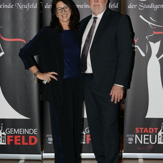 Ein Mann und eine Frau stehen eng nebeneinander vor einem Banner mit der Aufschrift 'Stadt Neufeld'. Die Frau trägt ein blaues Oberteil und schwarze Hosen, der Mann einen schwarzen Anzug. Beide lächeln.