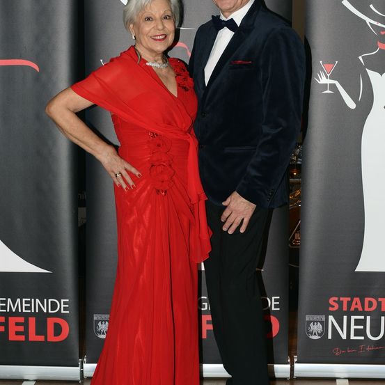 Ein Mann und eine Frau stehen lächelnd bei einer Veranstaltung, mit einem Hintergrund, der eine stilisierte Frau mit einem Glas Wein und den Worten 'Stadt Neu' zeigt.