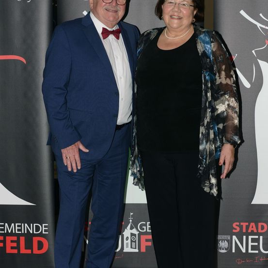 Ein Mann und eine Frau stehen eng beieinander und lächeln für ein Foto bei einer Veranstaltung. Der Mann trägt einen blauen Anzug mit einer roten Fliege. Die Frau trägt ein schwarzes Oberteil mit einer gemusterten Jacke. Hinter ihnen ist ein Hintergrund mit Text.