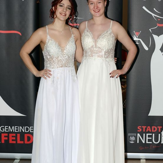 Zwei Frauen in weißen Kleidern stehen nebeneinander und posieren für ein Foto vor einem schwarzen Hintergrund mit rotem Text.