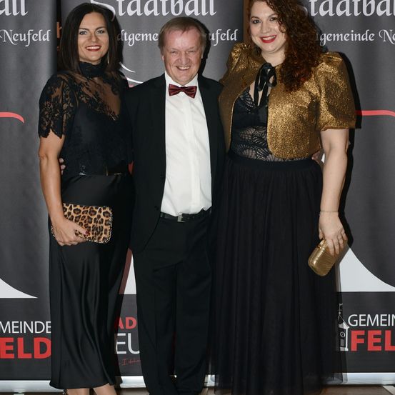 Drei Menschen, zwei Frauen und ein Mann, feierlich gekleidet, stehen zusammen vor einer Kulisse. Die Frauen lächeln.