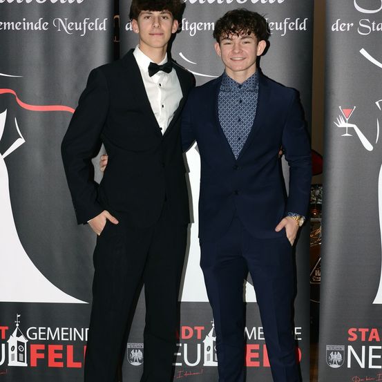 Zwei junge Männer in formeller Kleidung stehen nebeneinander und lächeln für die Kamera. Einer trägt einen schwarzen Frack mit einer Fliege, der andere einen marineblauen Anzug mit einem gemusterten Hemd. Sie stehen vor einem Hintergrund mit Text und Logos.