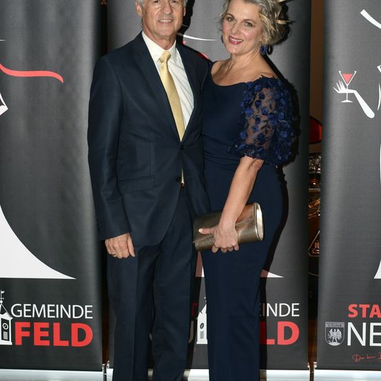 Ein Mann und eine Frau, beide formell gekleidet, stehen lächelnd vor einem Banner. Der Mann trägt einen dunklen Anzug und eine Krawatte, während die Frau ein marineblaues Kleid trägt.