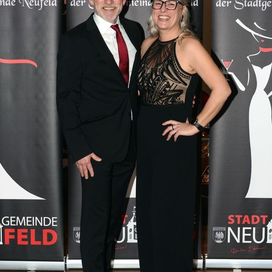 Ein Mann und eine Frau stehen lächelnd zusammen bei einer Veranstaltung. Der Mann trägt einen schwarzen Anzug und eine rote Krawatte. Die Frau trägt ein schwarzes Kleid mit einem weißen und schwarzen Design. Sie stehen vor einem Hintergrund, auf dem 'Stadt Neufeld' steht.