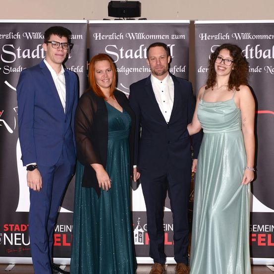 Vier Personen posieren für ein Foto vor einem Hintergrund mit der Aufschrift 'Herzlich Willkommen zum Stadt'. Der Mann links trägt einen blauen Anzug, die Frau in der Mitte trägt ein grünes Kleid und die Frau rechts trägt ein grünes Kleid und eine Brille. Der Mann in der Mitte trägt einen schwarzen Anzug und eine Fliege.