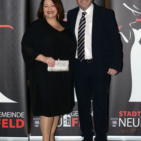 Eine lächelnde Frau in einem schwarzen Kleid und ein Mann in einem schwarzen Anzug und Krawatte stehen vor einem Banner mit dem Gesicht einer Frau und dem Wort 'Stadt Neufeld'.