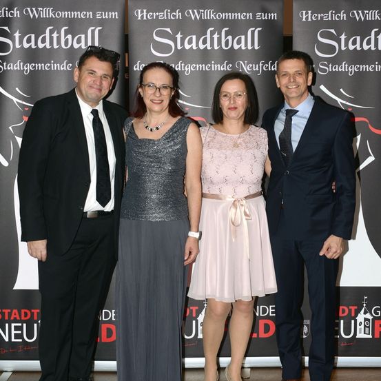 Vier formell gekleidete Personen stehen lächelnd zusammen vor einem Banner mit 'Stadtball' in weißen Buchstaben. Sie scheinen sich bei einer Veranstaltung zu befinden.
