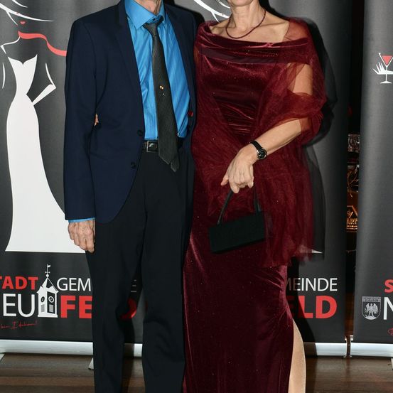 Ein älteres Paar, schick gekleidet, steht bei einer Veranstaltung. Der Mann trägt einen dunklen Anzug mit blauem Hemd und Krawatte, während die Frau ein rotes Kleid mit einer schwarzen Handtasche trägt. Sie stehen vor einem Banner mit rotem und weißem Text.