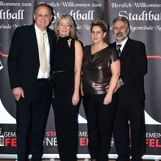 Vier Menschen stehen bei einer Veranstaltung zusammen und lächeln. Zwei Männer und zwei Frauen. Der Hintergrund hat roten und schwarzen Text.