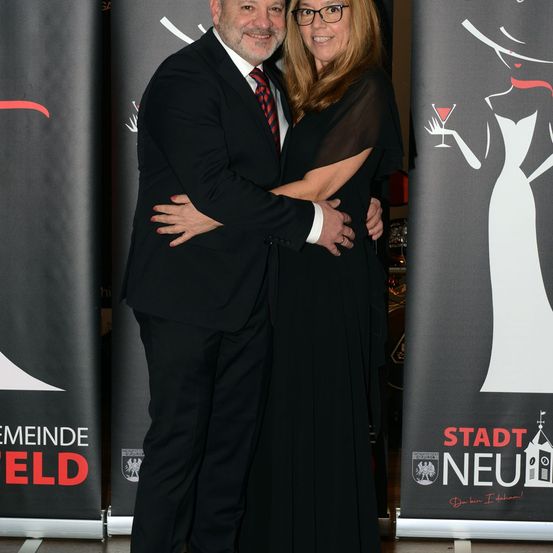 Ein älteres Paar posiert für ein Foto, in formeller Kleidung. Der Mann trägt eine Krawatte und die Frau trägt eine Brille. Sie stehen eng beieinander und haben ihre Arme umeinander. Sie stehen vor einem Plakat.