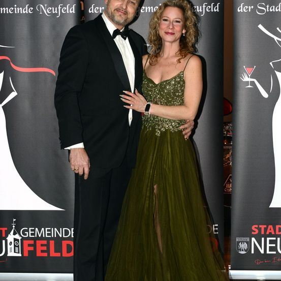 Ein Mann und eine Frau stehen bei einer Veranstaltung eng zusammen, in formeller Kleidung. Die Frau trägt ein grünes Kleid mit hohem Schlitz, und der Mann einen schwarzen Anzug. Sie stehen vor einem Hintergrund mit Logos und Text.