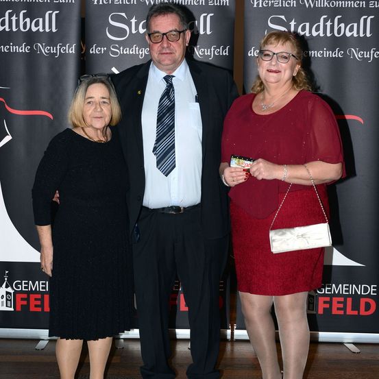 Drei Personen stehen zusammen auf einer Bühne mit einem Banner im Hintergrund. Die Frau auf der linken Seite trägt ein schwarzes Kleid, während die zentrale Figur, ein Mann, einen Anzug und eine Krawatte trägt. Die Frau auf der rechten Seite hält eine kleine weiße Tasche.